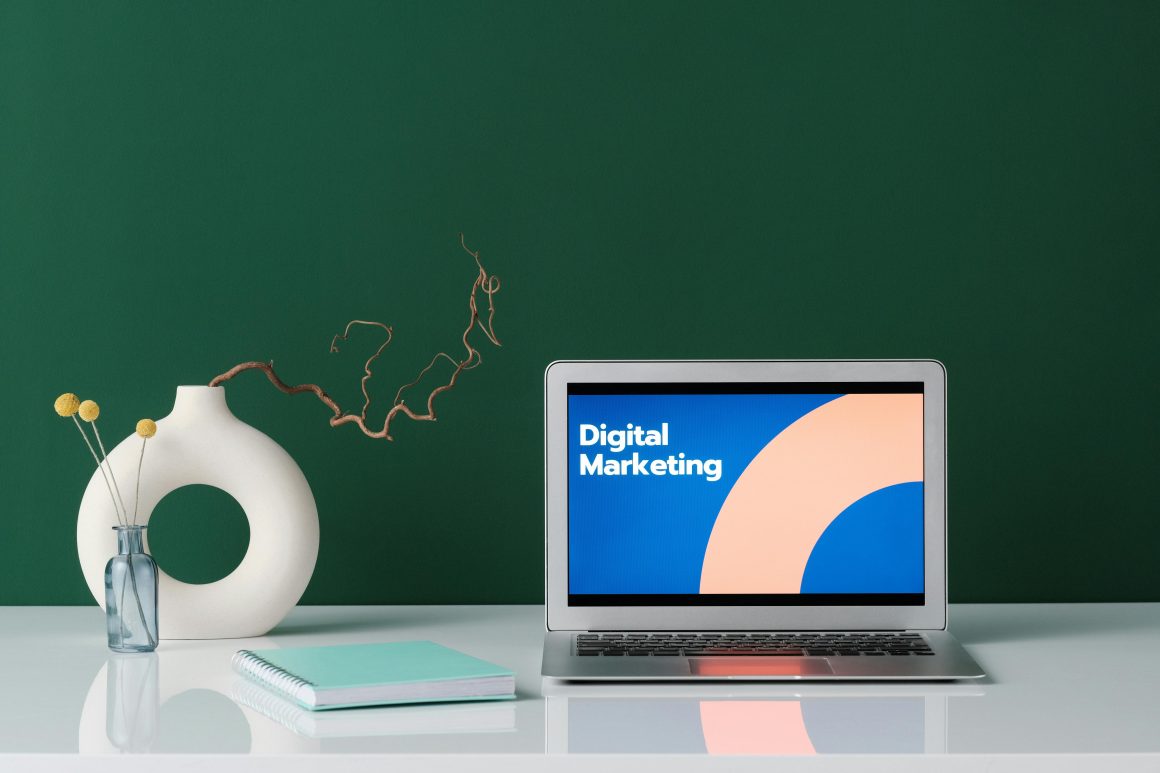 Laptop displaying 'Digital Marketing' on a white desk with a vase, notebook, and decorative elements against a green background.