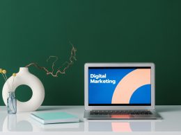 Laptop displaying 'Digital Marketing' on a white desk with a vase, notebook, and decorative elements against a green background.