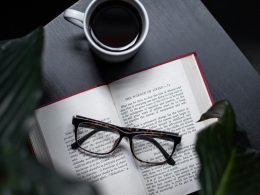 Open self-help book with glasses resting on its pages, next to a cup of black coffee on a dark wooden table, surrounded by green leaves, creating a calm and reflective atmosphere.