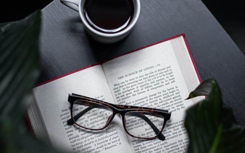 Open self-help book with glasses resting on its pages, next to a cup of black coffee on a dark wooden table, surrounded by green leaves, creating a calm and reflective atmosphere.