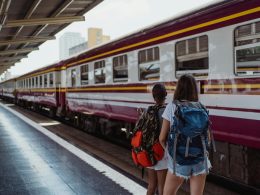 Two backpackers standing on a train platform, facing a maroon and white train. Ready to board, representing smart ways to travel on a budget."