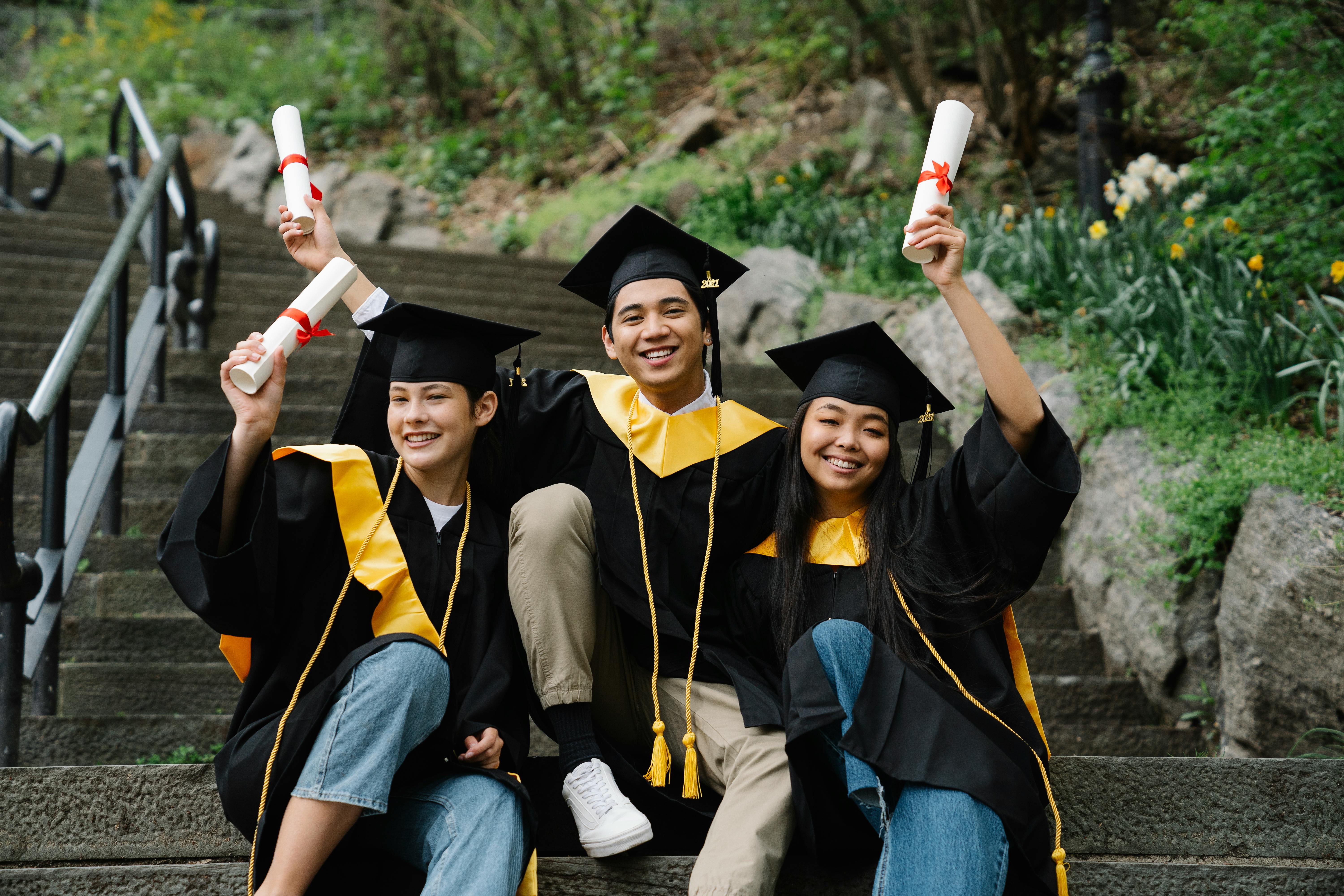 Three happy graduates in black and gold gowns celebrating on outdoor stairs, holding diplomas—symbolizing bright careers after school.