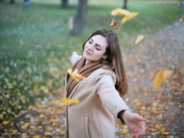 A young woman in a beige coat with arms outstretched, embracing the falling autumn leaves with a peaceful smile, symbolizing the path to joy and freedom.