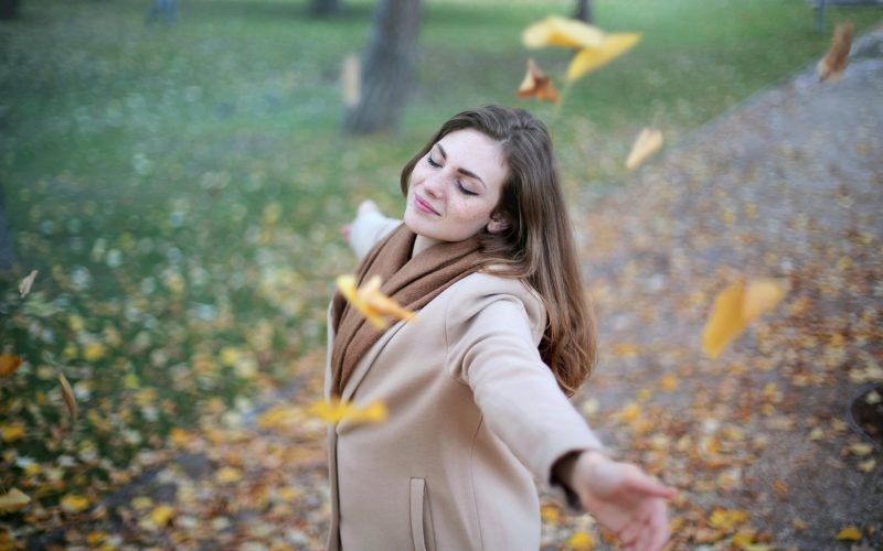 A young woman in a beige coat with arms outstretched, embracing the falling autumn leaves with a peaceful smile, symbolizing the path to joy and freedom.